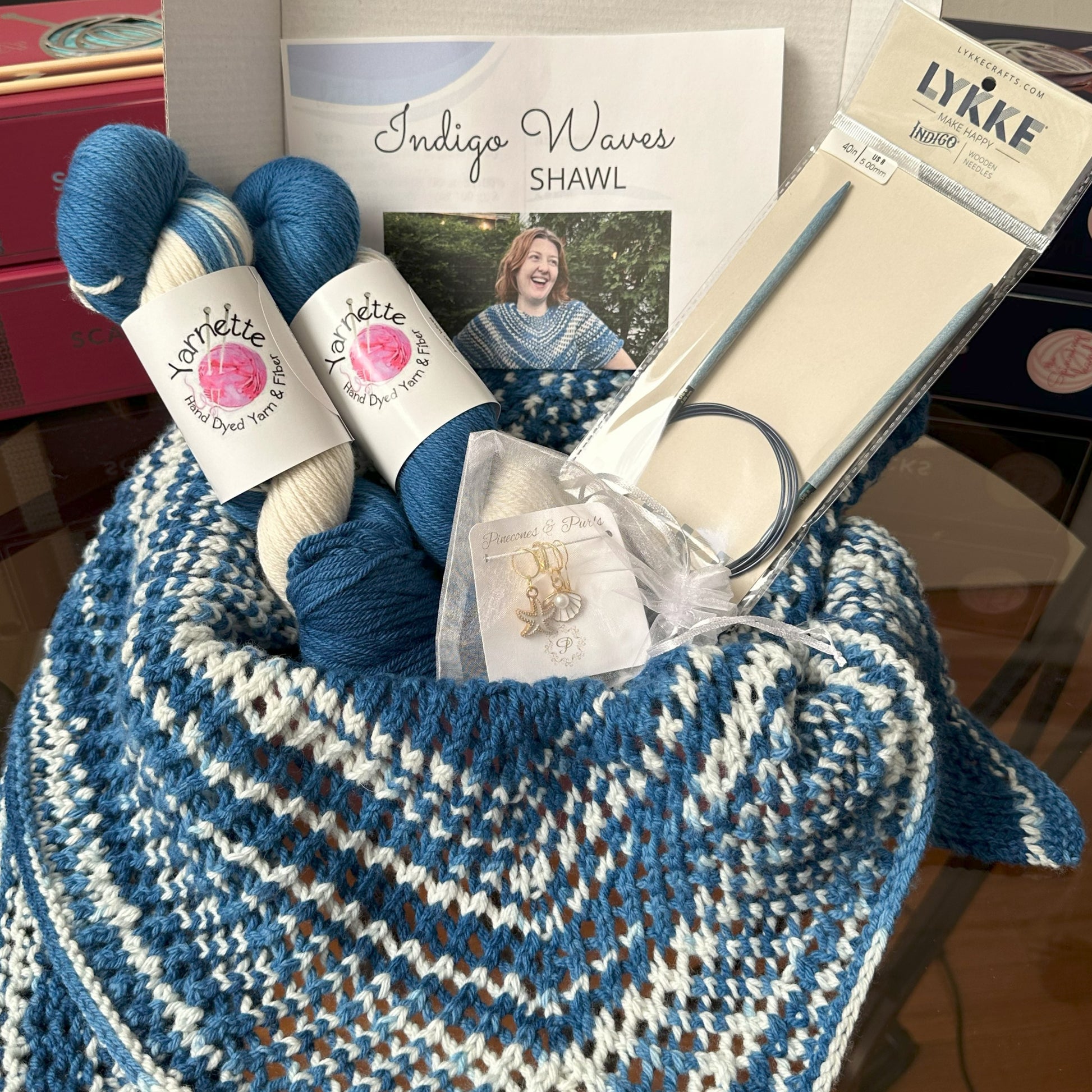 Knitting kit with yarn, needles, and a completed shawl on a wooden surface.