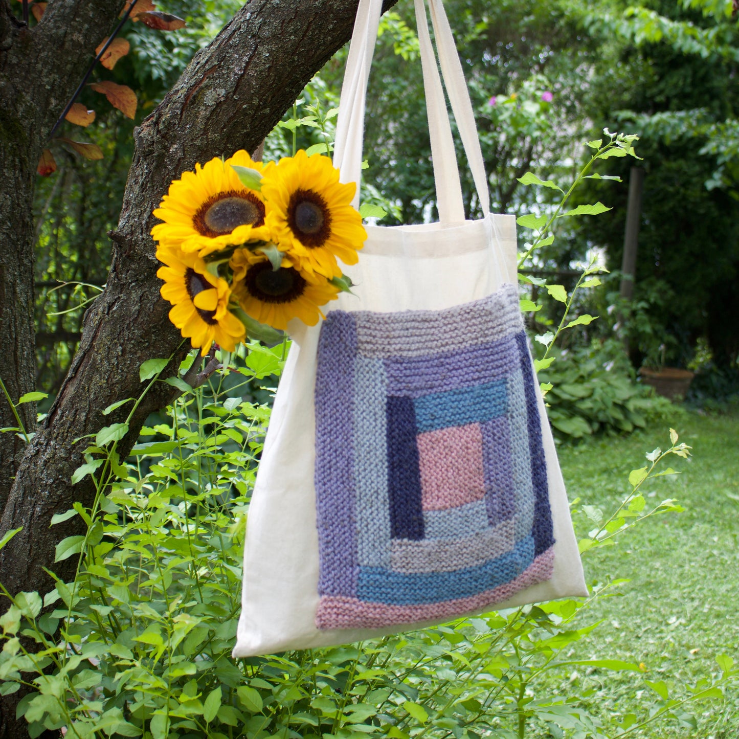 A cream canvas tote bag with a colorful hand-knit geometric patchwork design in shades of blue, purple, and pink, hanging from a tree branch in a lush green garden. Two yellow sunflowers are tucked into the side of the bag.