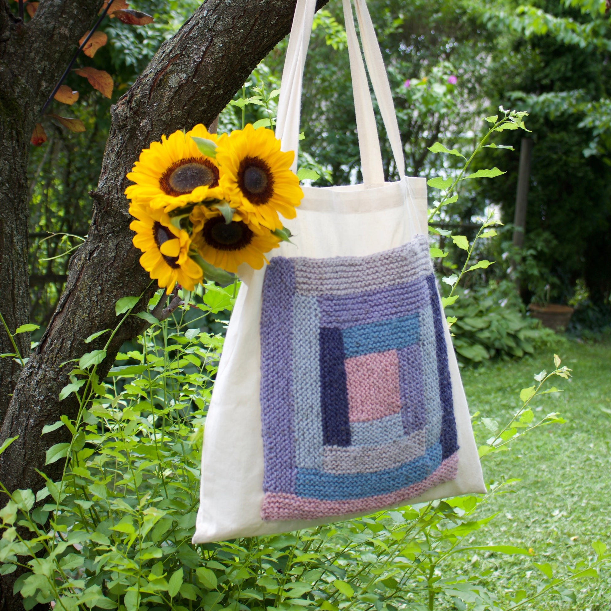 A cream canvas tote bag with a colorful hand-knit geometric patchwork design in shades of blue, purple, and pink, hanging from a tree branch in a lush green garden. Two yellow sunflowers are tucked into the side of the bag.