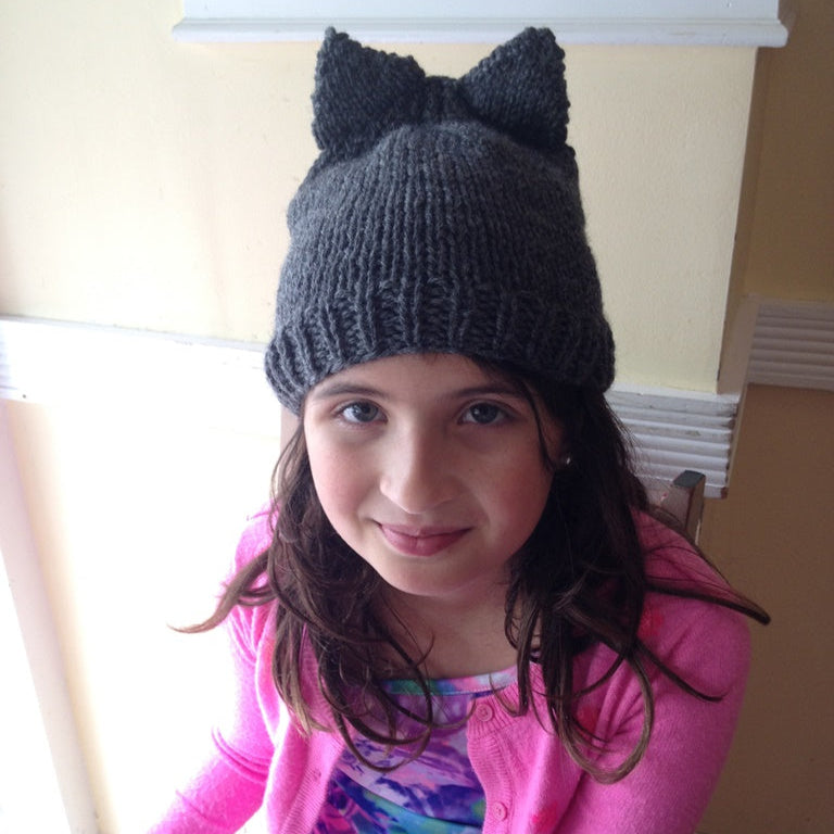 Young girl wearing a dark gray knit hat with cat ears indoors.
